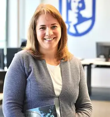 Frau mit braunen Haaren lächelt in die Kamera. Im Hintergrund ist ein Büro zu sehen mit einem großen ROLAND Assistance Logo.