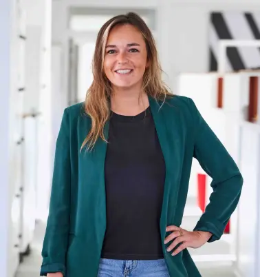 Eine Frau mit blonden Haaren steht in einem Büroflur. Sie trägt ein graues Oberteil und hat eine Hand in der Taille. Sie lächelt.