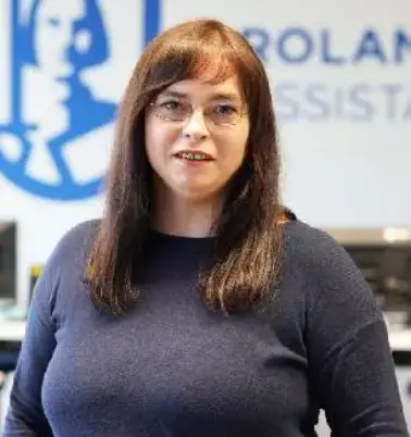 Frau mit braunen Haaren und Brille lächelt in die Kamera. Im Hintergrund ist ein Büro zu sehen mit einem großen ROLAND Assistance Logo.