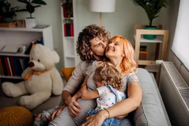 Ein junges Paar sitz mit Kleinkind au feinem Sofa. Sie kuscheln und lachen.
