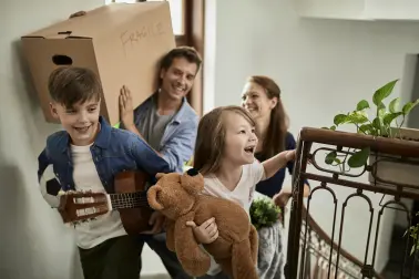 Famile zieht um und lacht. Sie steigen in einem Hausflur die Treppe herauf. Die zwei Kinder tragen Spielsachen, die Eltern tragen Kisten.