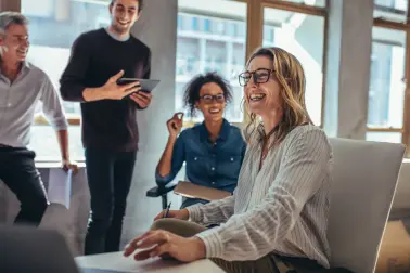 Frau mit Brille sitzt an einem Tisch in einem Meetingraum. Vor ihr steht ein Laptop. Neben ihr sitzt eine junge Frau. Zwei Männer stehen neben ihnen. Alle lachen. 