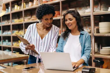 Zwei junge Frauen stehen in einem Keramikladen. Sie schauen sich gemeinsam etwas auf einem Laptop an. Auf dem Tisch vor ihnen liegen ein Smartphone und ein EC-Lesegerät. Eine der Frauen hat ein Klemmbrett in der Hand. Beide lachen.
