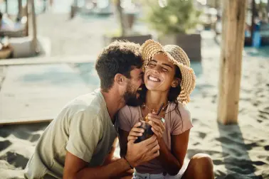 Junge Frau mit Strohhut sitzt an Strand und hält einen Cocktail in der Hand. Neben ihr sitzt ein junger Mann und umarmt sie. Er hat eine Hand um den Cocktail. Beide lachen ausgelassen.