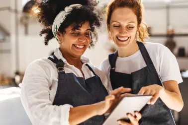 Zwei Frauen mit Schürzen schauen zusammen auf ein Tablet und lächeln.