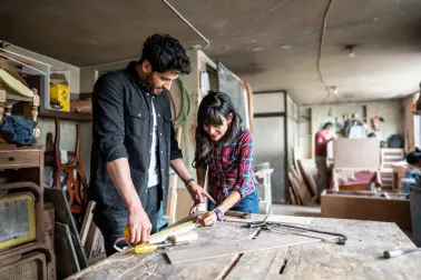 Junge Frau und junger Mann stehen in einer Schreinerwerkstatt an einem Holztisch. Sie messen mit einem Maßband ein Stück Holz ab. Auf dem Tisch liegen eine Säge, eine Feile und ein Schraubenzieher. Beide lächeln.