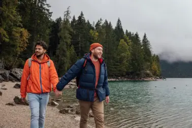 Zwei junge Männer halten sich an den Händen und spazieren an einem Wald entlang. Neben ihnen ist ein See. Beide tragen Wanderkleidung und Rucksäcke. Sie lächeln. 