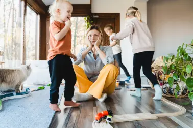 Eine junge Mutter sitzt auf dem Boden neben ihren Kindern. Sie hat den Kopf auf die Hände gestützt und schaut betrübt. Um sie herum liegen viele Spielsachen und die Kinder laufen umher.