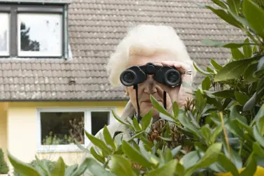 Eine Rentnerin steht im Garten und schaut durch ein Fernglas.
