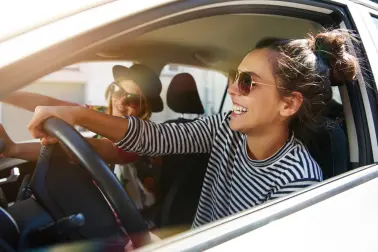 Zwei junge Frauen fahren Auto, tragen Sonnenbrillen und lachen.