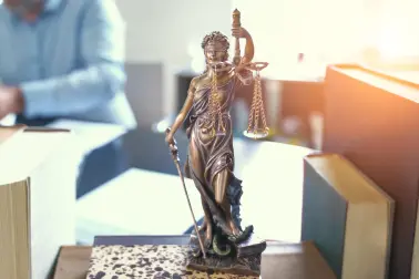 Auf einem Schreibtisch steht auf einem Buch eine Bronze Statue. Diese Statue zeigt Justitia. Sie hat die Augen verbunden, trägt in einer Hand ein Schwert und in der anderen eine Waage mit zwei Waagschalen. An dem Schreibtisch sitzt jemand im Hemd und arbeitet an Akten. Auf dem Tisch stehen weitere dicke Bücher.