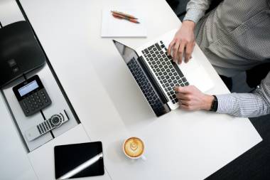 Person in gestreiftem Hemd sitzt an Schreibtisch und arbeitet an Macbook. Auf dem Schreibtisch liegen ein Notizblock mit Stift, ein Taschenrechner, ein Ipad und eine Fernbedienung.