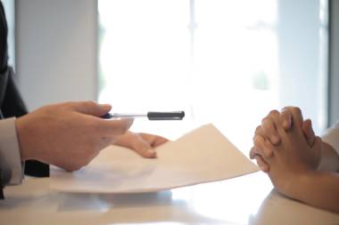 Zwei Personen sitzen sich gegenüber an einem Tisch. Eine Person hält Unterlagen und einen Kugelschreiber in der Hand. Die andere Person hat die Hände gefaltet.