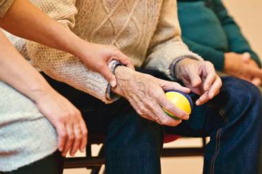Ein Senior sitzt und hält in der Hand einen kleinen Ball. Neben ihm sitzt eine andere Person und hat die Hand auf seinem Arm.