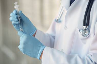 Person im weißen Kitte und blauen Einweghandschuhen zieht eine Spritze für eine Impfung auf. Die Person trägt ein Stethoskop um den Hals.