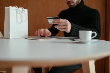 Mann sitzt an Tisch, hält Kreditkarte in der Hand und bedient sein Tablet.