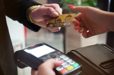 Person mit goldener Uhr am Handgelenk reicht goldene VISA Karte an eine andere Person mit pinken Fingernägeln. Diese hat ein Kartenlesegerät in der anderen Hand.