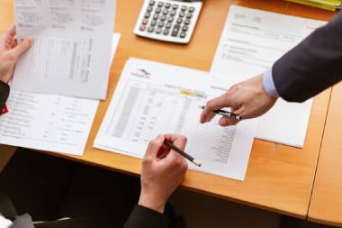 Auf einem Tisch liegen Abrechnungen und Tabellen. Zwei Personen arbeiten an diesen, sie halten jeweils einen Stift in der Hand.