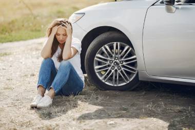 Junge Frau sitzt auf Boden vor einem stehenden Auto. Sie hat die Ellbogen auf die Knie aufgestützt und hat die Hände am Kopf.