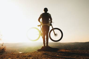 Person mit Rucksack steht auf Berggipfel. Die Person hält ein Fahrrad in der Hand.