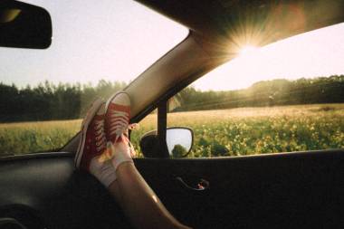 Person mit roten Schuhen hat diese auf das Amaturenbrett eines Autos gelegt. Dieses Auto fährt an einem blühenden Feld vorbei.