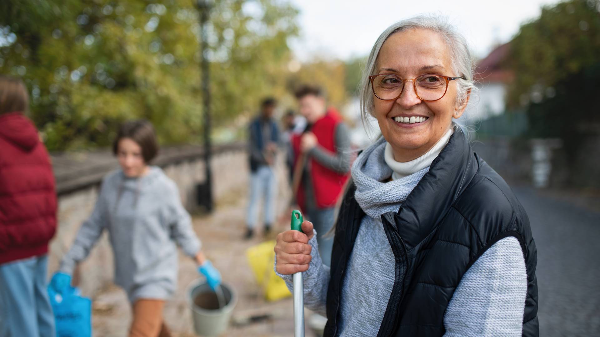 Ältere Frau mit Brille lächelt. Im Hintergrund sammeln Menschen Müll auf.