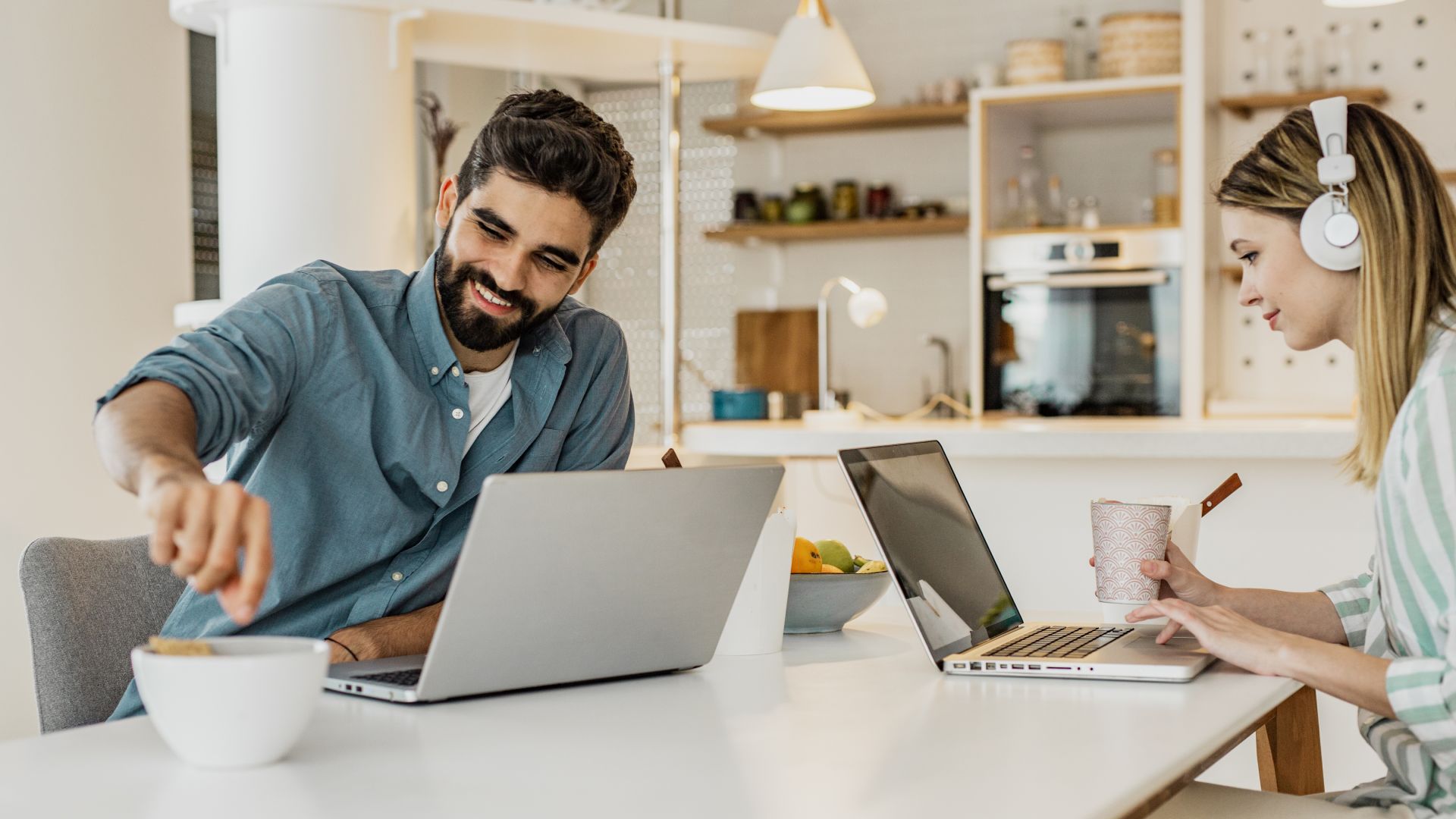 Junge Frau und junger Mann sitzen zusammen an Tisch in Wohnungsküche. Die Frau trägt Kopfhörer. Beide haben Laptops vor sich.