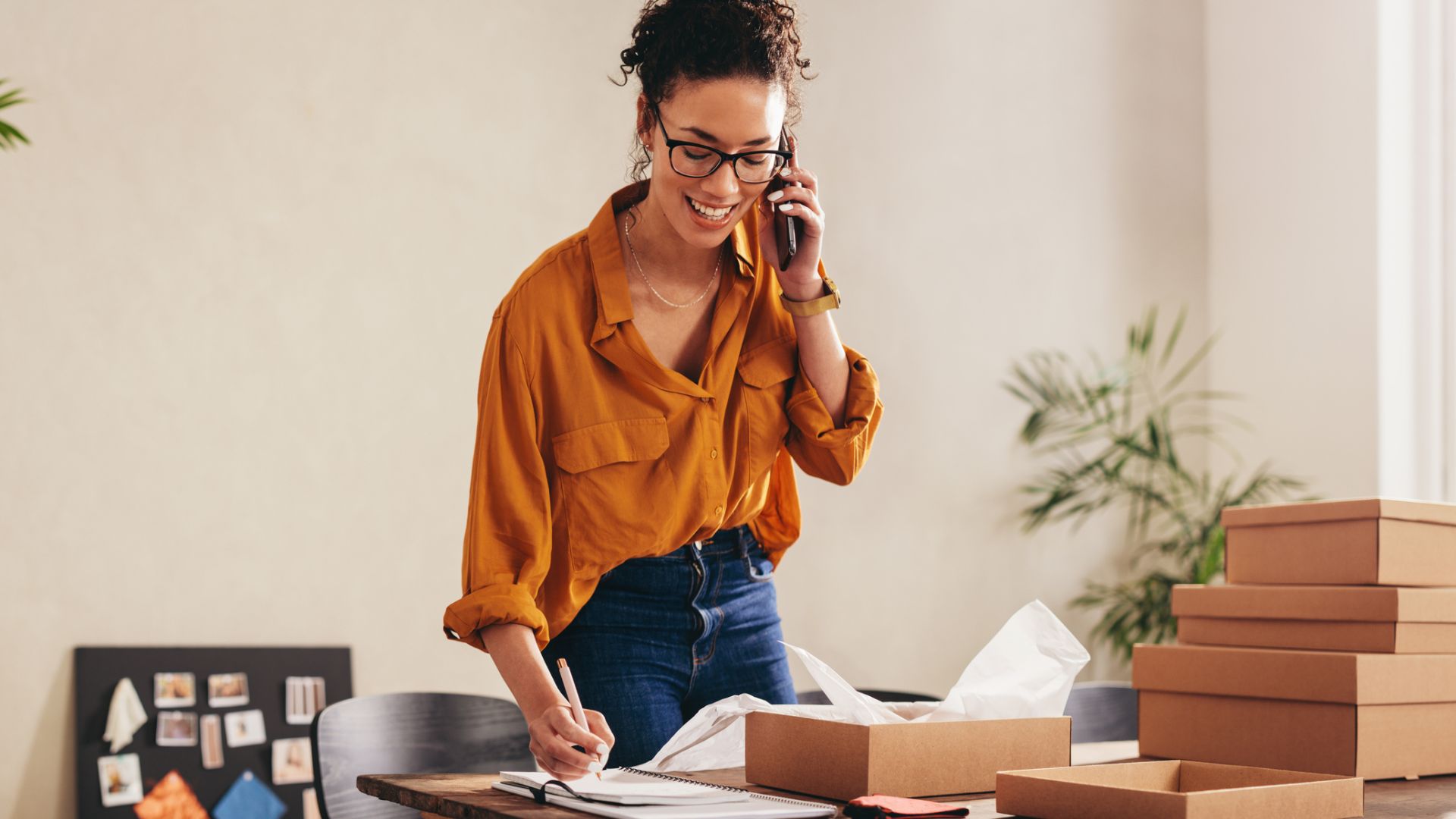 Frau steht vor einem Tisch und telefoniert. Mit der rechten Hand macht sie schriftliche Notizen. Auf dem Tisch stehen mehrere Kartons.