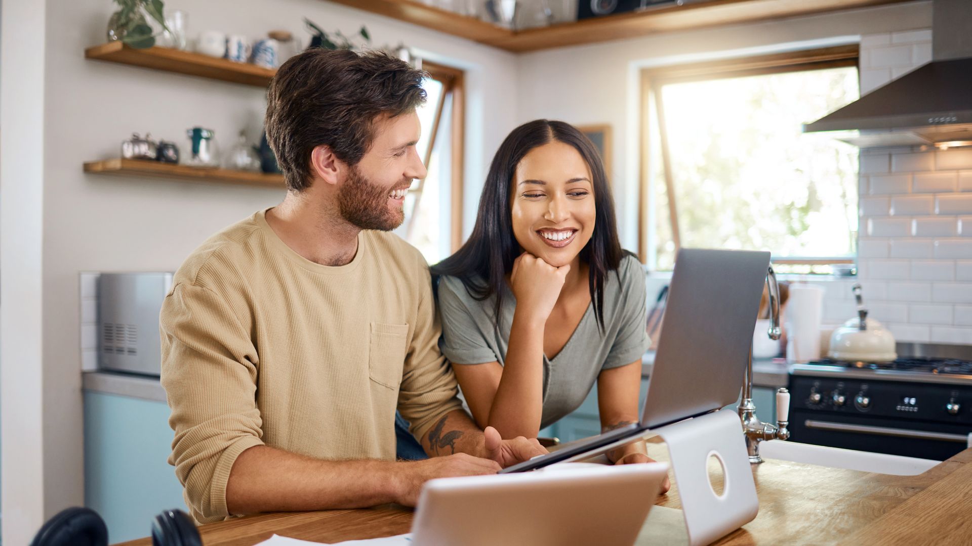 Junges Paar steht in Wohnungsküche und schaut sich etwas auf einem Laptop an. Beide lachen. Auf dem Tisch steht ein Tablet neben unterschiedlichen Dokumenten.