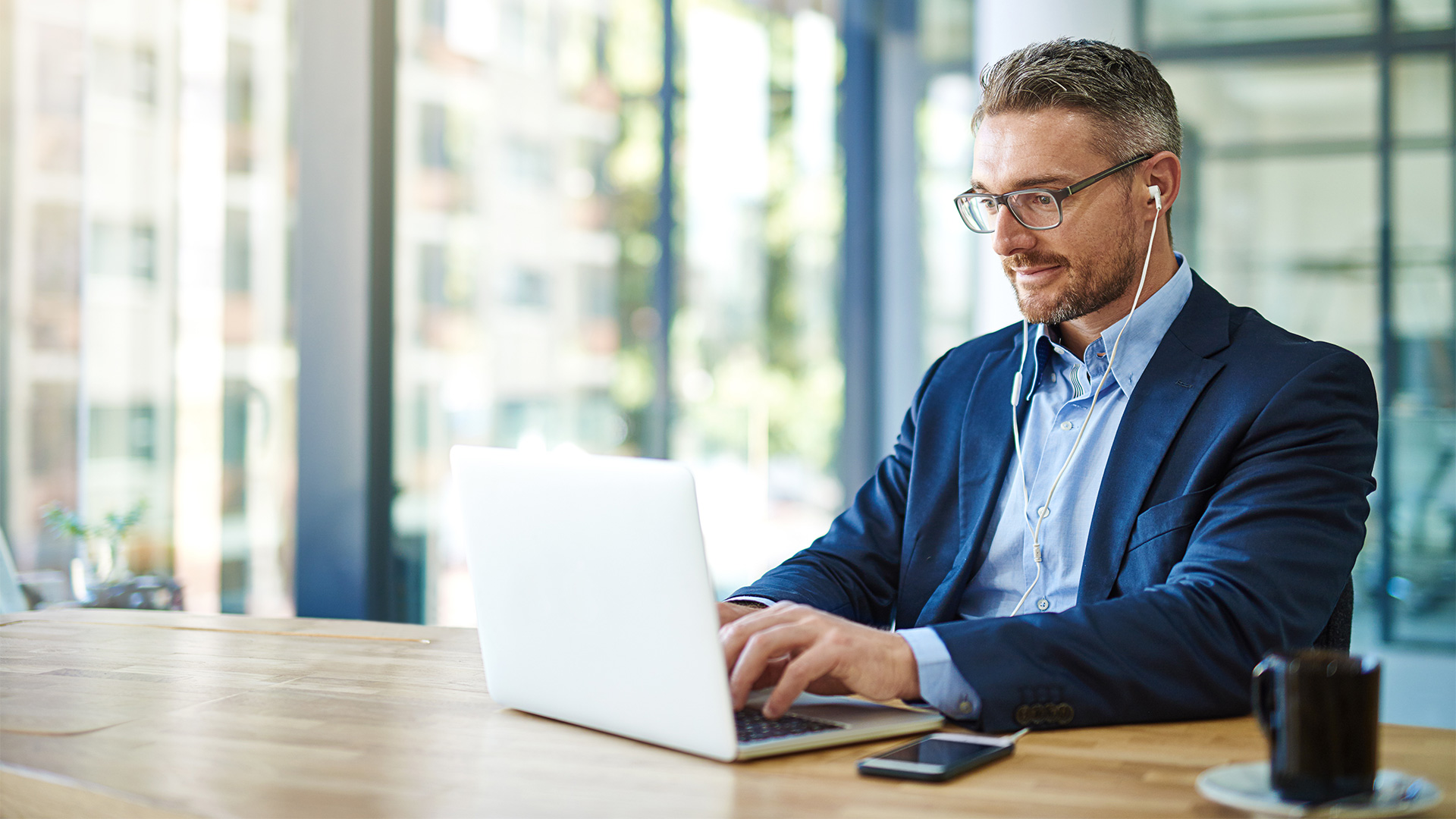 Mann in Anzug sitzt an einem Tisch. Er schreibt etwas auf seinem Laptop. Er hat weiße Kabelkopfhörer in den Ohren, welche in sein Smartphone gesteckt sind. Das Smartphone liegt neben dem Laptop auf dem Tisch.