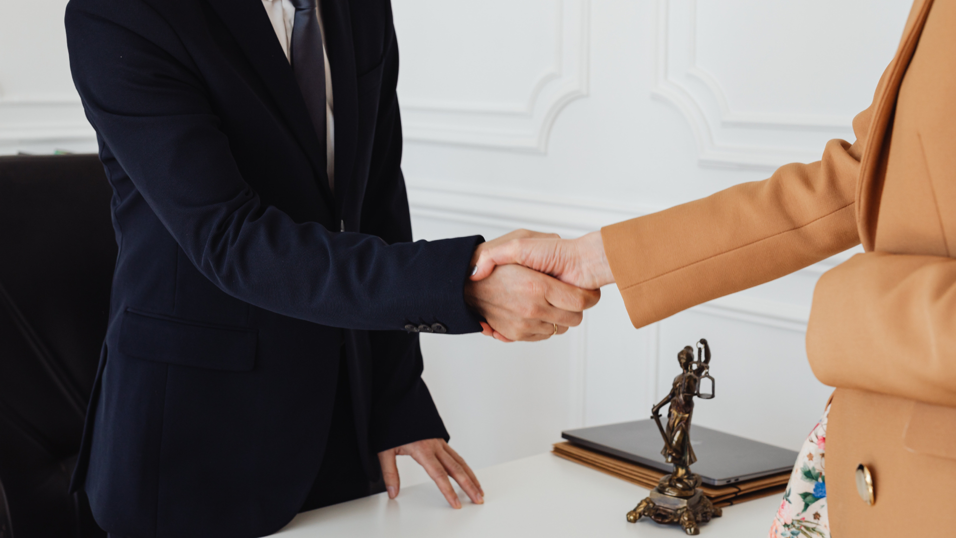 Auf einem Schreibtisch steht eine Bronze Statue. Diese Statue zeigt Justitia. Sie hat die Augen verbunden, trägt in einer Hand ein Schwert und in der anderen eine Waage mit zwei Waagschalen. Über den Schreibtisch hinweg geben sich zwei Personen die Hand. Eine Person trägt einen Anzug. Die andere Person ein Kleid mit Blumen und einen Mantel.