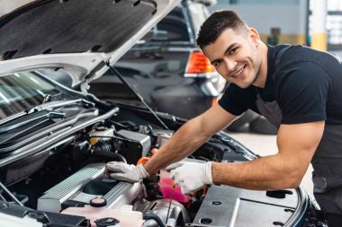 Junger Mann arbeitet an offener Motorhaube. Er trägt Arbeitskleidung und Handschuhe.