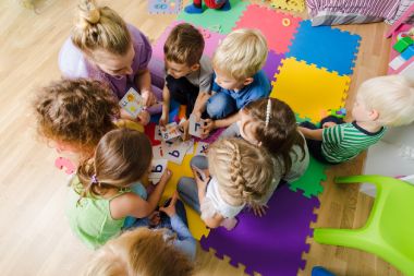 Eine Erzieherin sitzt auf dem Boden auf einem bunten Teppich in einer KiTa. Um sie herum sitzen 8 Kinder. Die Kinder lernen mit der Erzieherin Buchstaben mit bunten Karten. Um sie herum sind weitere bunte Spielzeuge.
