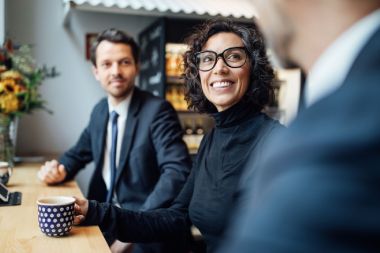 Frau sitzt am Tisch, hält Tasse in der Hand und lächelt. Sie spricht mit einem Mann im Anzug. Neben ihr sitzt ein weiterer Mann im Anzug, welcher den beiden zuhört.