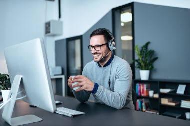 Mann mit Brille trägt Headset und sitzt an Schreibtisch. Er schaut auf einen Bildschirm und hält eine Kaffeetasse in der Hand. Er lacht.