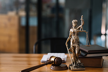 Statue der Justitia auf dem Schreibtisch. Die Statue hat die Augen verbunden, trägt in einer Hand ein Schwert und in der anderen eine Waage mit zwei Waagschalen.