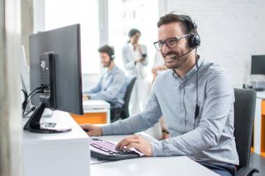 Junger Mann sitzt in Büro an Schreibtisch und telefoniert über ein Headset. Er macht die telefonische Rechtsberatung. Er lächelt, schaut auf dem Bildschirm vor ihm und bedient die Tastatur. Neben ihm sitzt eine weitere Person an einem Schreibtisch. Zwei weitere Personen stehen und unterhalten sich