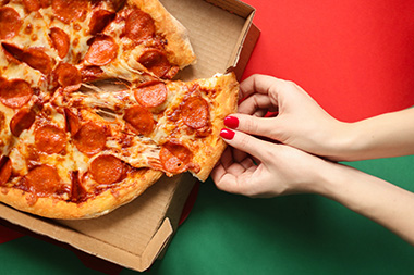 Person mit rotem Nagellack nimmt sich ein Stück Salami-Pizza aus einem Pizzakarton aus Pappe.