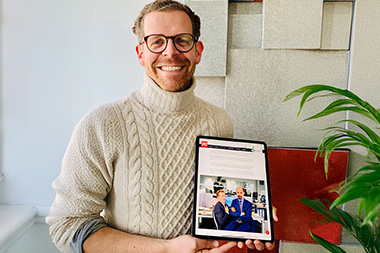 Tobias von Mäßenhausen hält ein iPad in der Hand. Er schaut nach vorne und lächelt. Das iPad zeigt einen Artikel über 